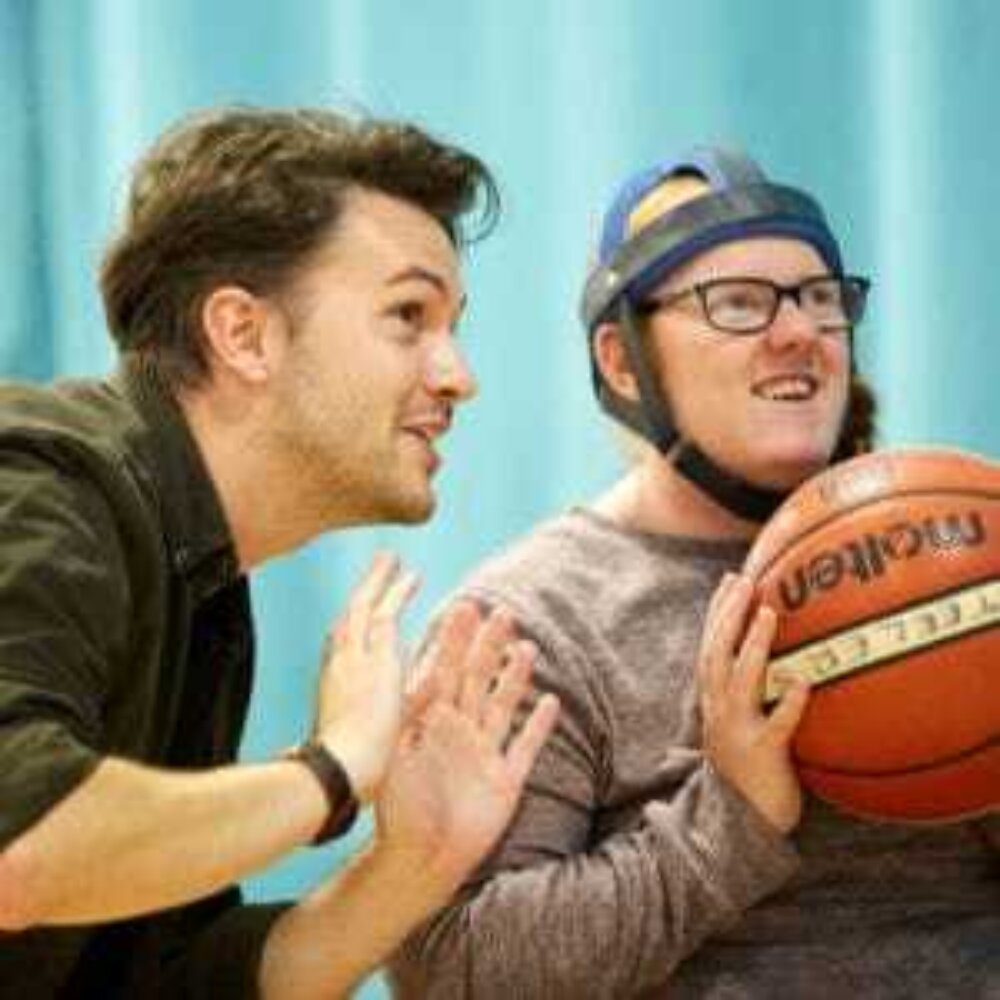 A young man is holding a basketball. He's sitting and about to throw it, and someone is next to him coaching him