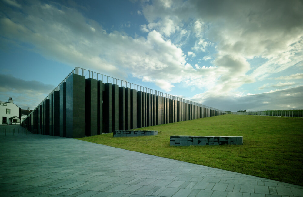 A large modern building with stone pillars. It is long and low under a vast cloudy sky