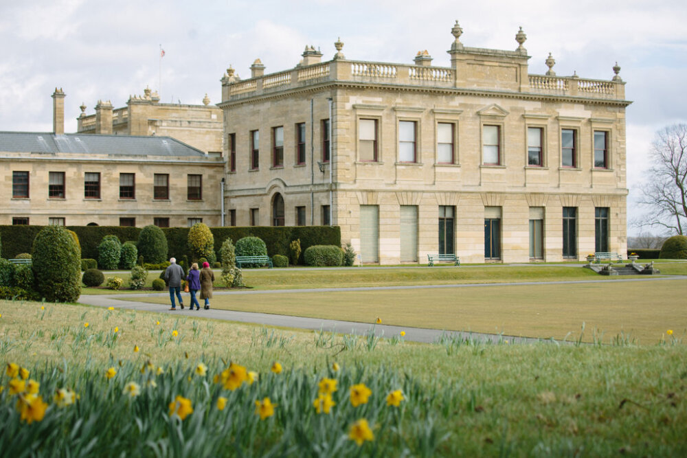 A grand stone mansion with manicured gardens in the foreground, featuring blooming yellow daffodils. Three people stroll on a path under a cloudy sky.