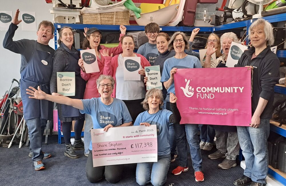Photo shows an excited group of volunteers inside Share Skipton's library of things, holding a giant cheque, and celebrating their grant of £117,398 from the National Lottery Community Fund. Behind them are a variety of items available for lending.
