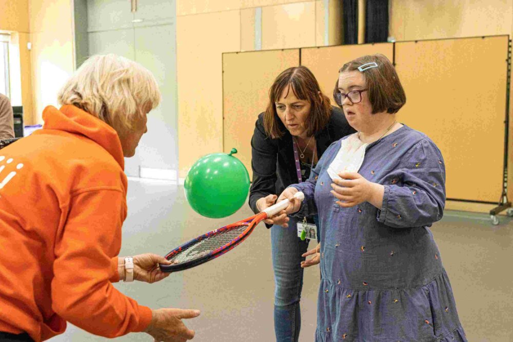 A female in a blue dress is holding a tennis racket and hitting a balloon in the air. her support worker is stood next to her encouraging her and the coach wearing and orange hoodie is helping her.