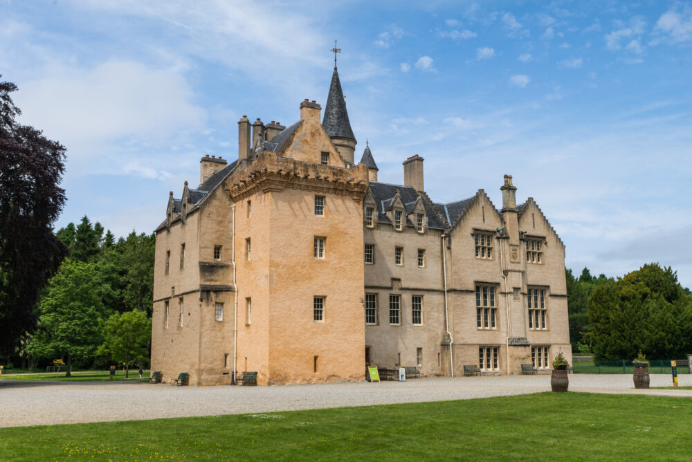 A historic stone castle with turrets, steep roofs, and tall chimneys, set beside a lawn under a bright blue sky.