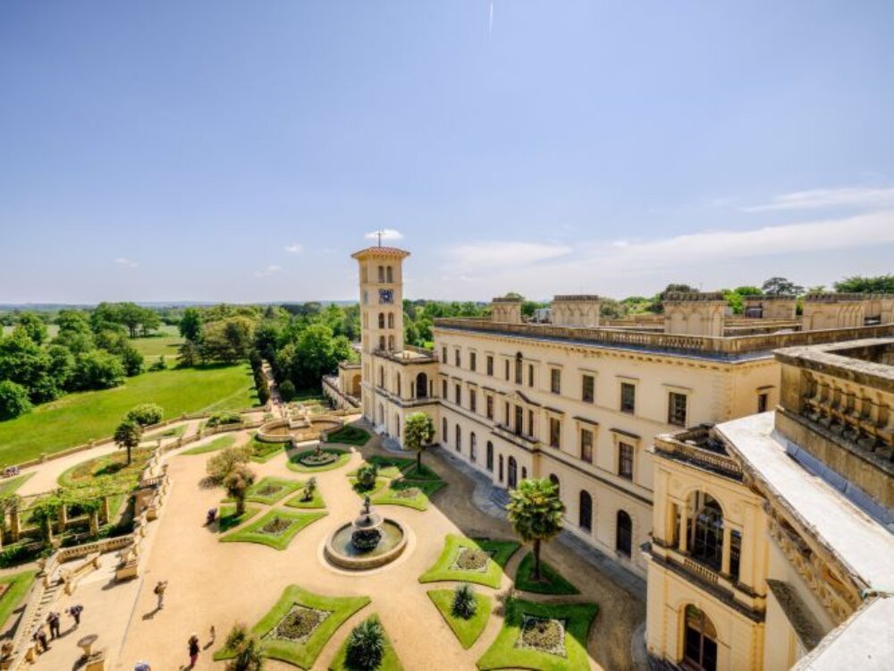 A grand historic building with a tall, square tower overlooks a manicured garden featuring a central fountain, surrounded by lush green fields under a clear blue sky.