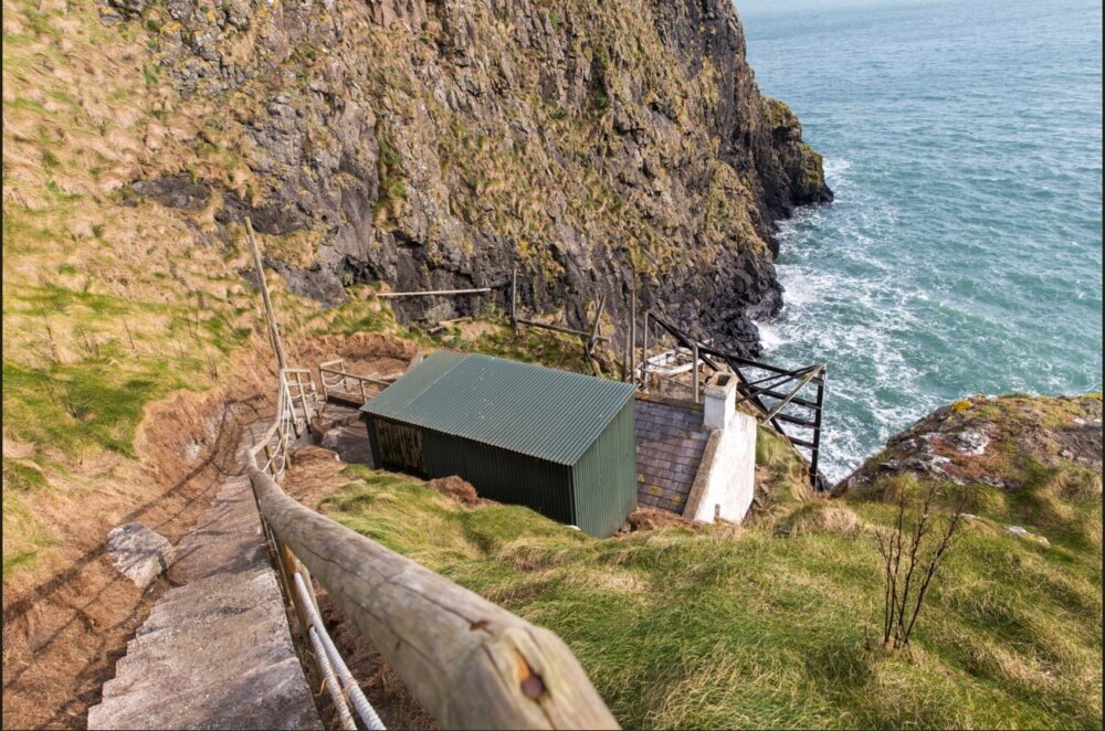 A small dark building with a tin roof nestled in the cliffs overlooking the sea