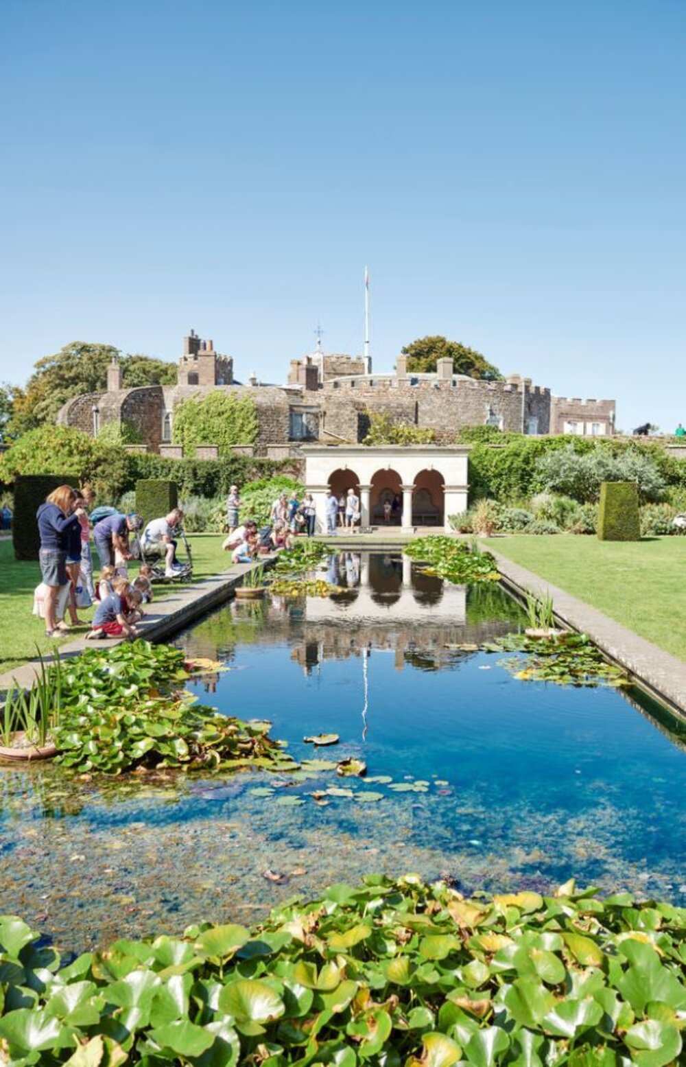 People gather around a reflective pond in a lush garden, overlooked by an old stone castle. The scene is lively and relaxed, on a clear day.